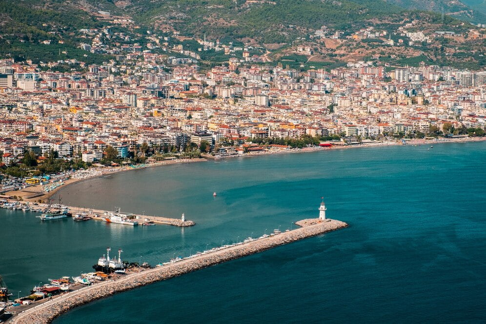 Aerial view of Alanya, a coastal resort town with residential buildings and a prominent lighthouse extending into the blue sea.
